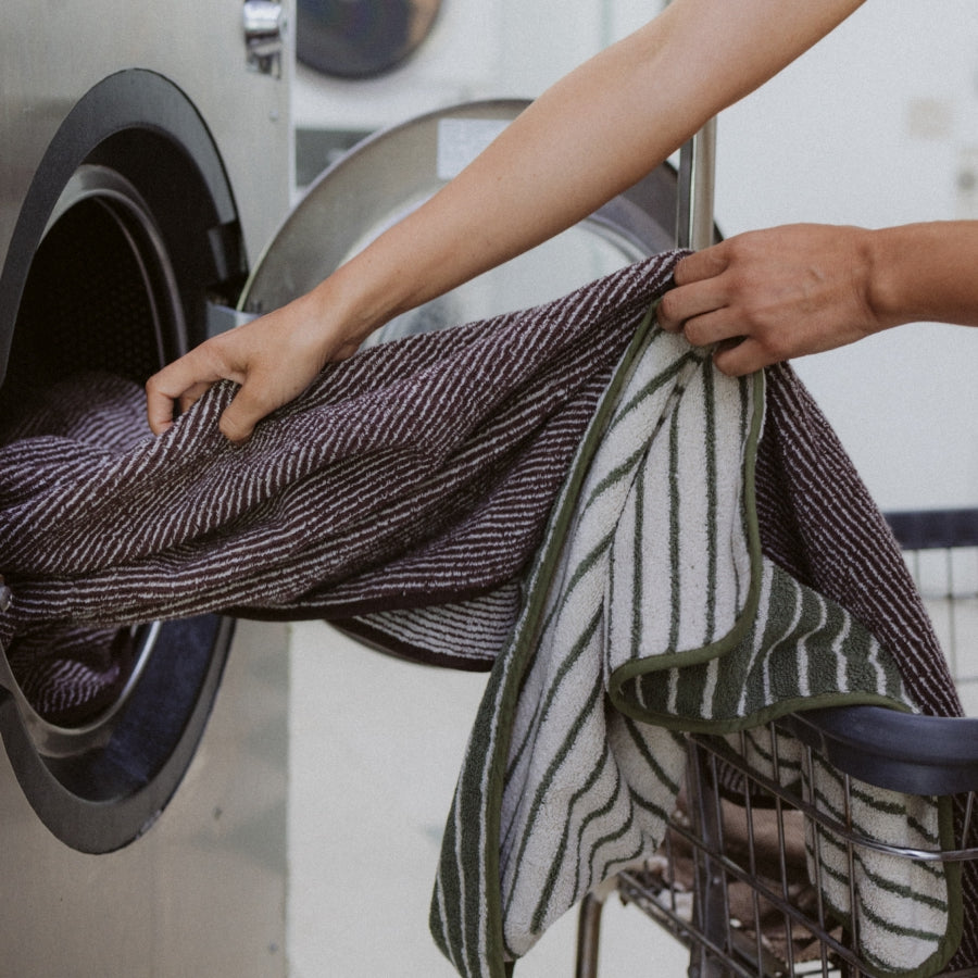 A close-up shot of a person's hands transferring a striped towel from a washing machine into a metal laundry basket. 