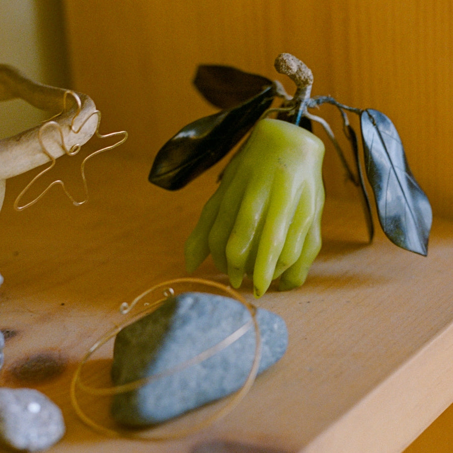 A green hand-shaped candle with dark leaves on a wooden surface, surrounded by small decorative stones and jewelry pieces.