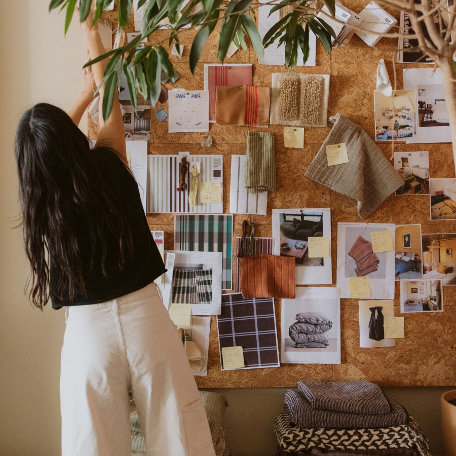 A woman stands in front of a corkboard wall, reaching toward hanging greenery. The board is covered with fabric swatches, photos, notes, and design samples in earthy tones.