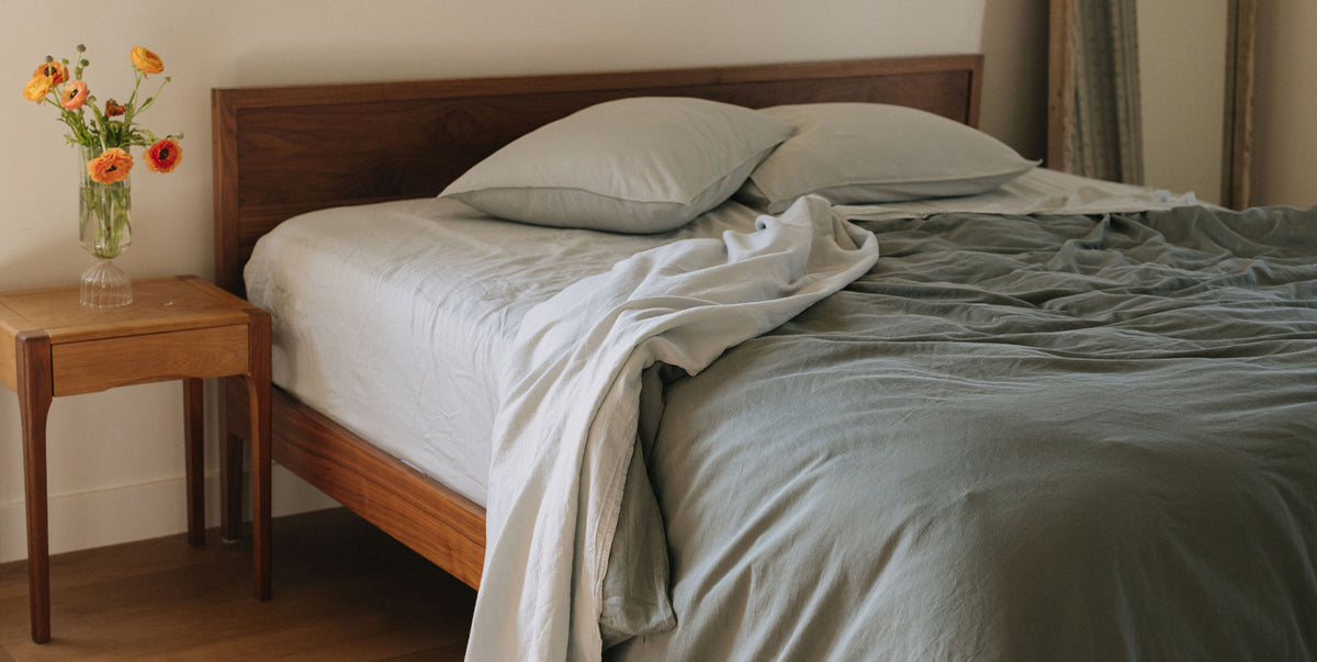 Bedroom with a wooden bed frame, light blue and green bedding, and a small table with flowers.