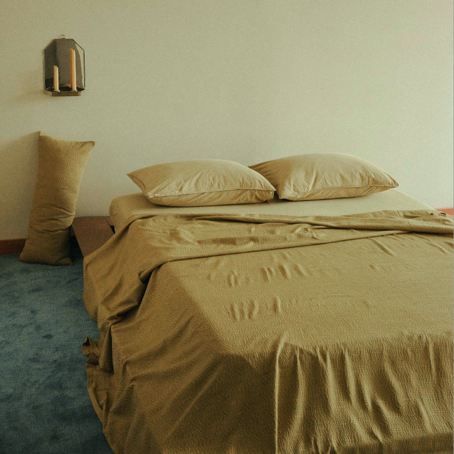 A bed dressed in muted olive-green bedding with matching pillows and a tall pillowcase, placed in a minimalist room with a light wall, blue carpet, and wall-mounted sconce.