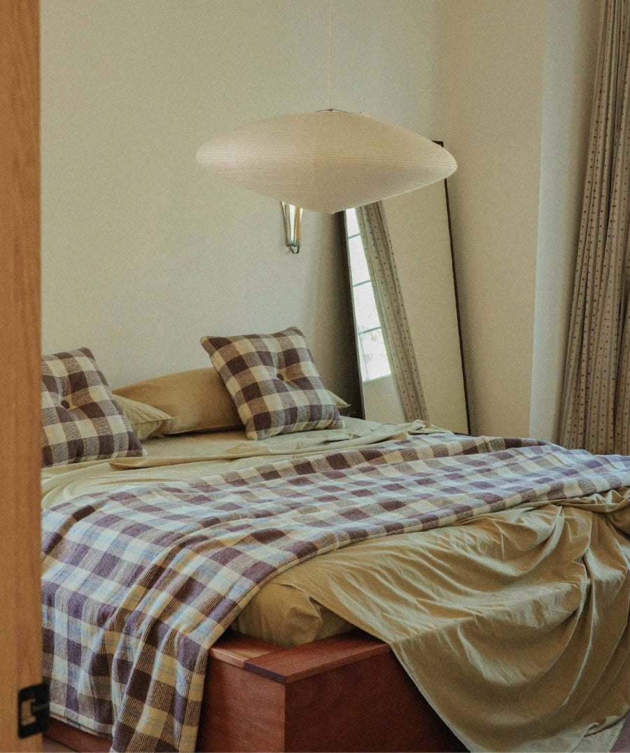A cozy bedroom scene with a bed dressed in light green sheets and a brown-and-beige plaid blanket and pillow shams, with a cream pendant light above and a tall mirror in the background.