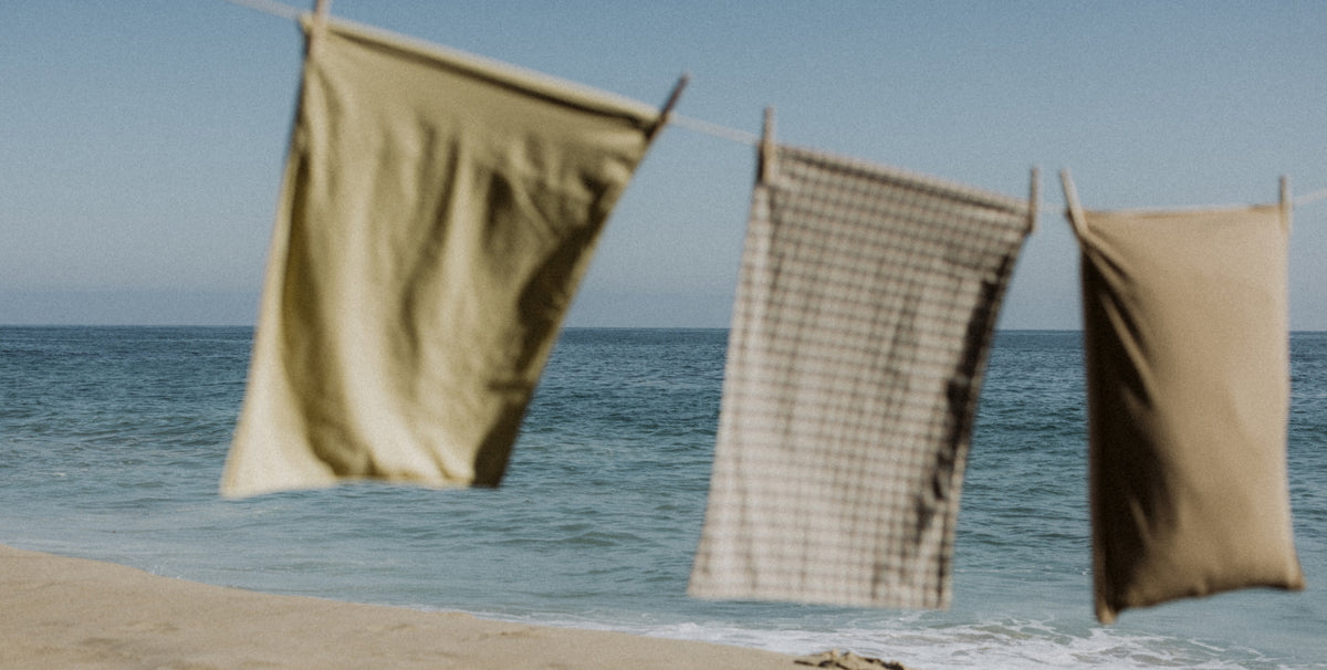 Three fabric cloths or towels hanging on a string line outdoors by the beach, with ocean water and pale sand in the background.