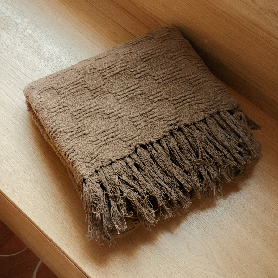 A folded brown textured throw blanket with fringed edges, placed on a light wood surface.