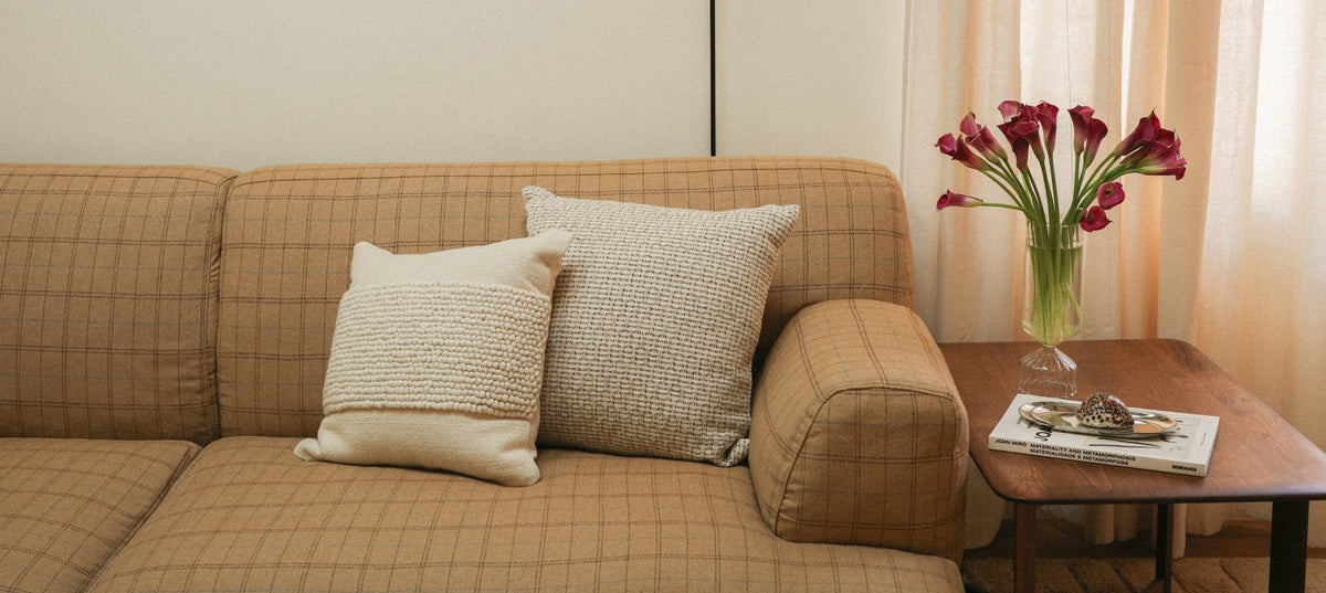A beige plaid sofa with two textured cream throw pillows, set in a cozy living room next to a wooden side table holding a vase of red flowers and a magazine.