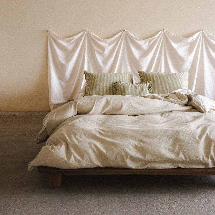 A minimalist bed styled with light green bedding and textured pillows, set against a draped cream fabric headboard in a warm, neutral-toned room.