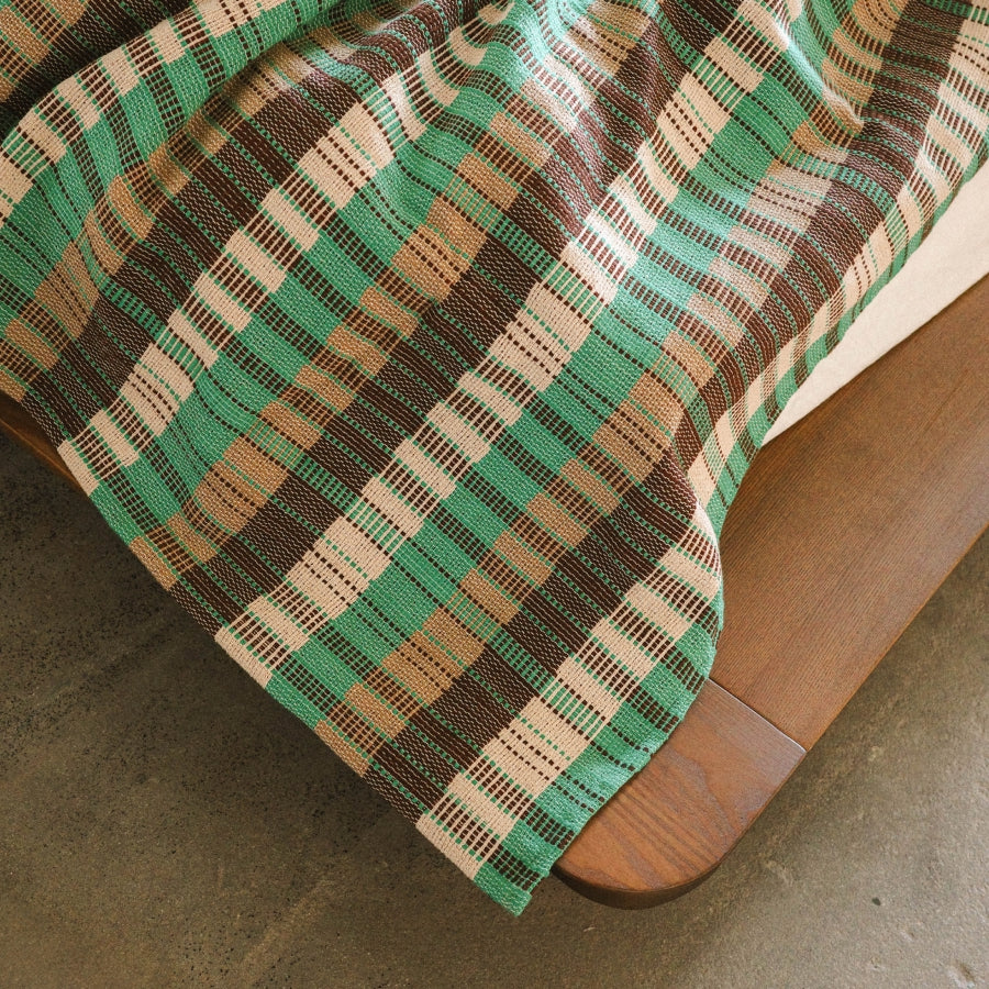 A close-up of a woven plaid blanket draped over the corner of a wooden bed frame, showing a pattern of green, brown, beige, and white stripes.