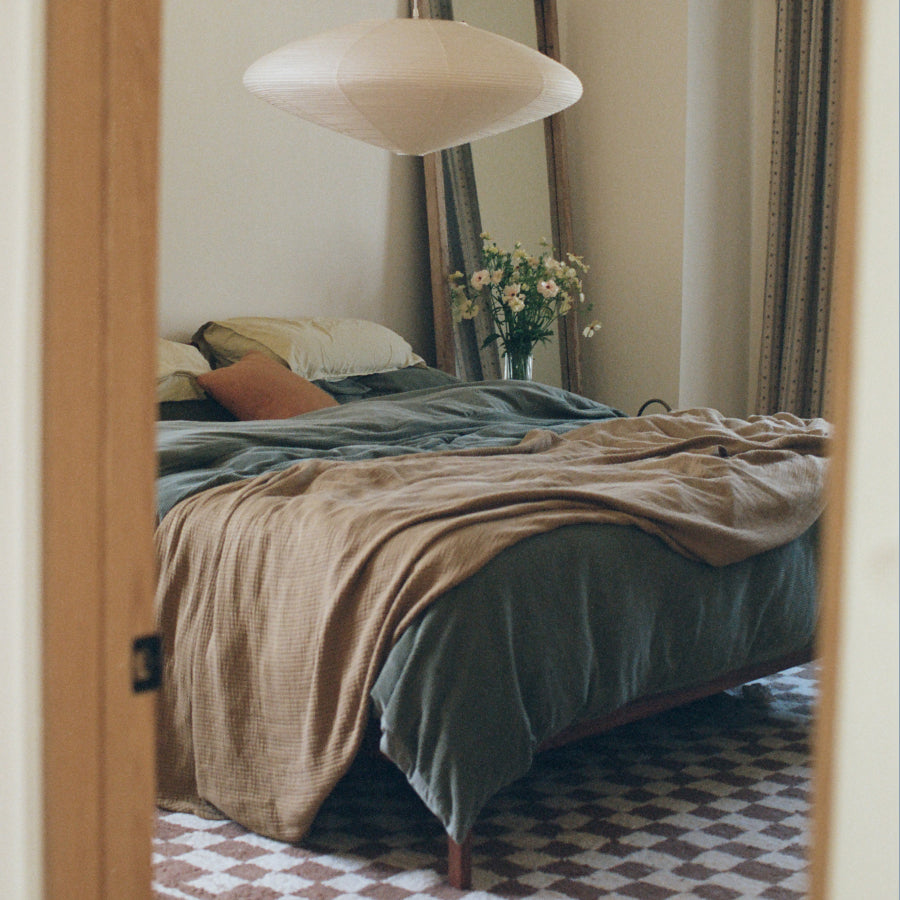 A cozy bedroom scene viewed through an open wooden doorway, featuring a bed dressed in muted sage green and tan textiles, a hanging paper lantern, curtains, and a vase of flowers.