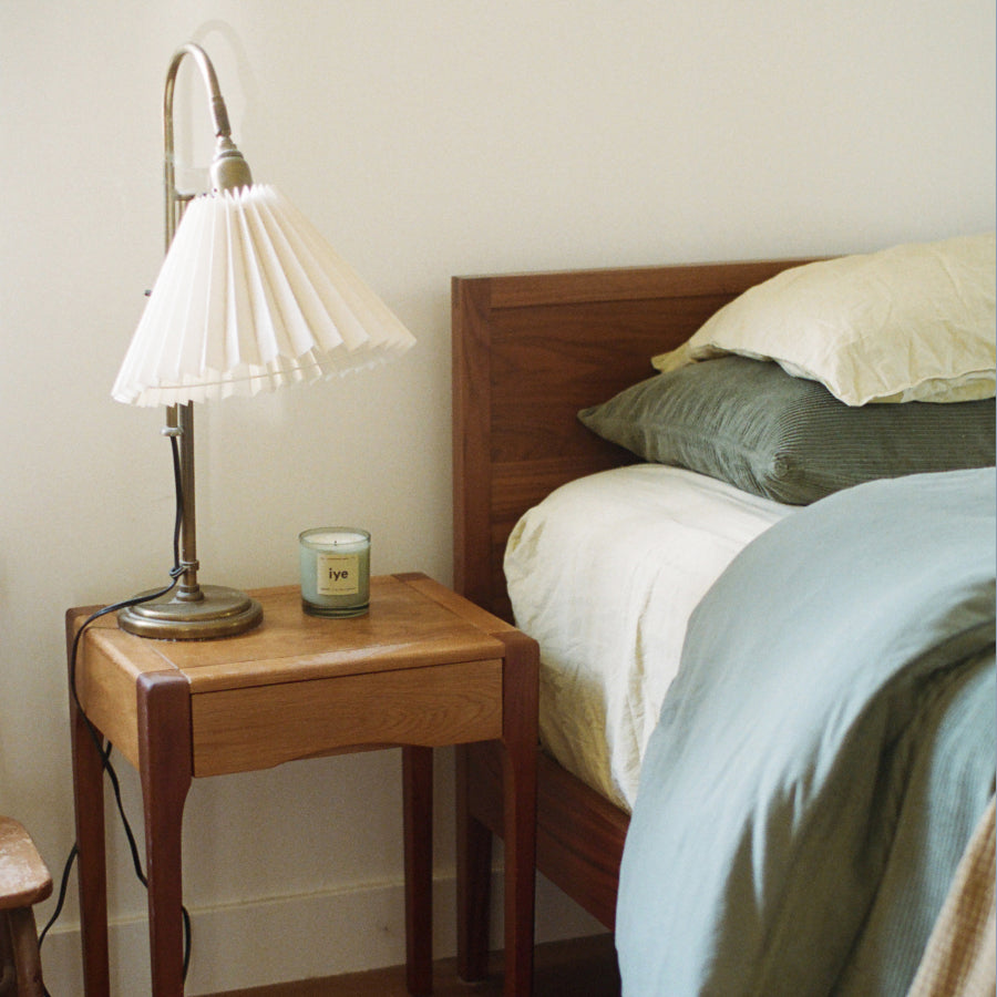 A mid-century modern table lamp with a pleated white shade and brushed metal base sits on a wooden nightstand next to a bed with cream bedding and a sage green pillow.