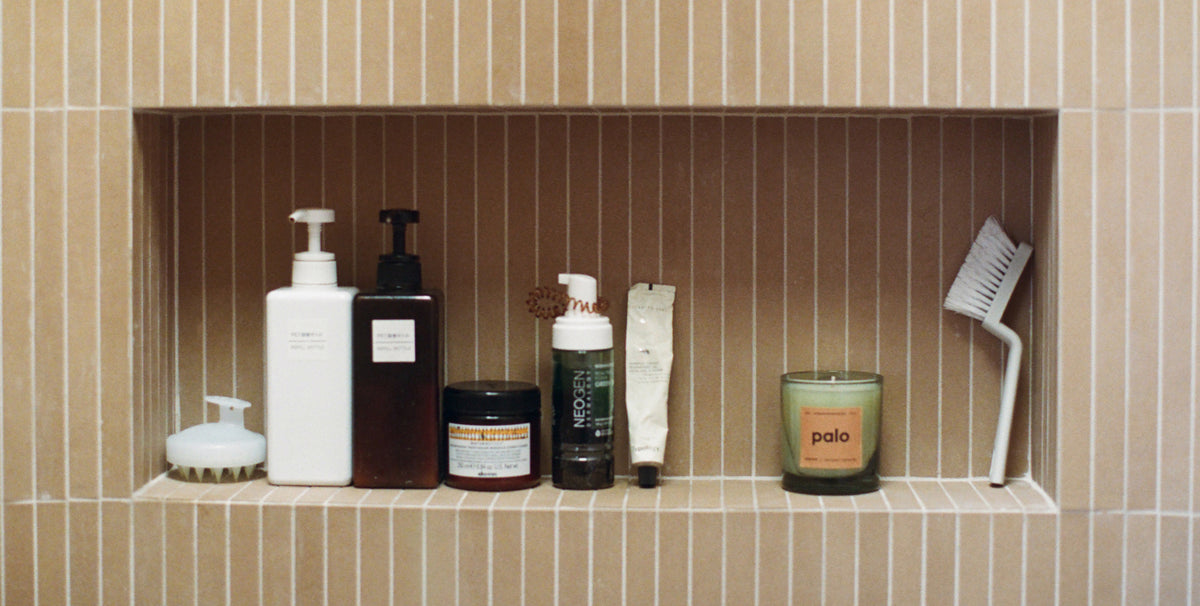 A collection of bathroom toiletries and accessories arranged on a recessed shelf against a beige tiled wall. Visible items include several pump bottles, a jar, a tube, a candle, and a toothbrush.