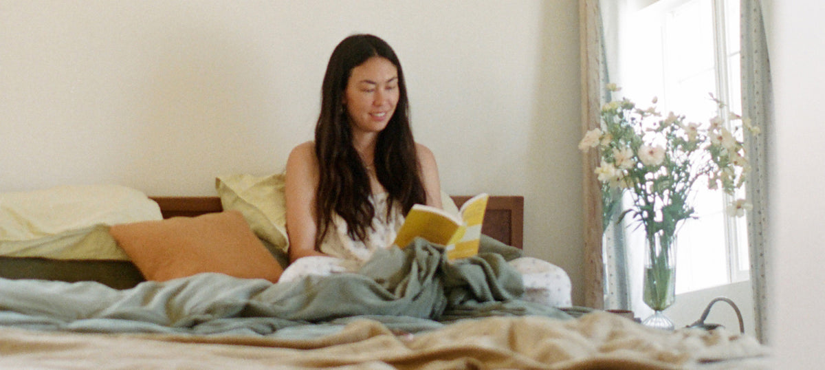 A woman sits in bed reading a yellow book, surrounded by green bedding, with a vase of flowers on a bedside table near a bright window.