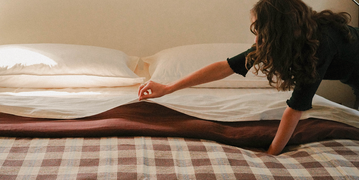 Person reaching towards a bed with white sheets and brown plaid blanket.