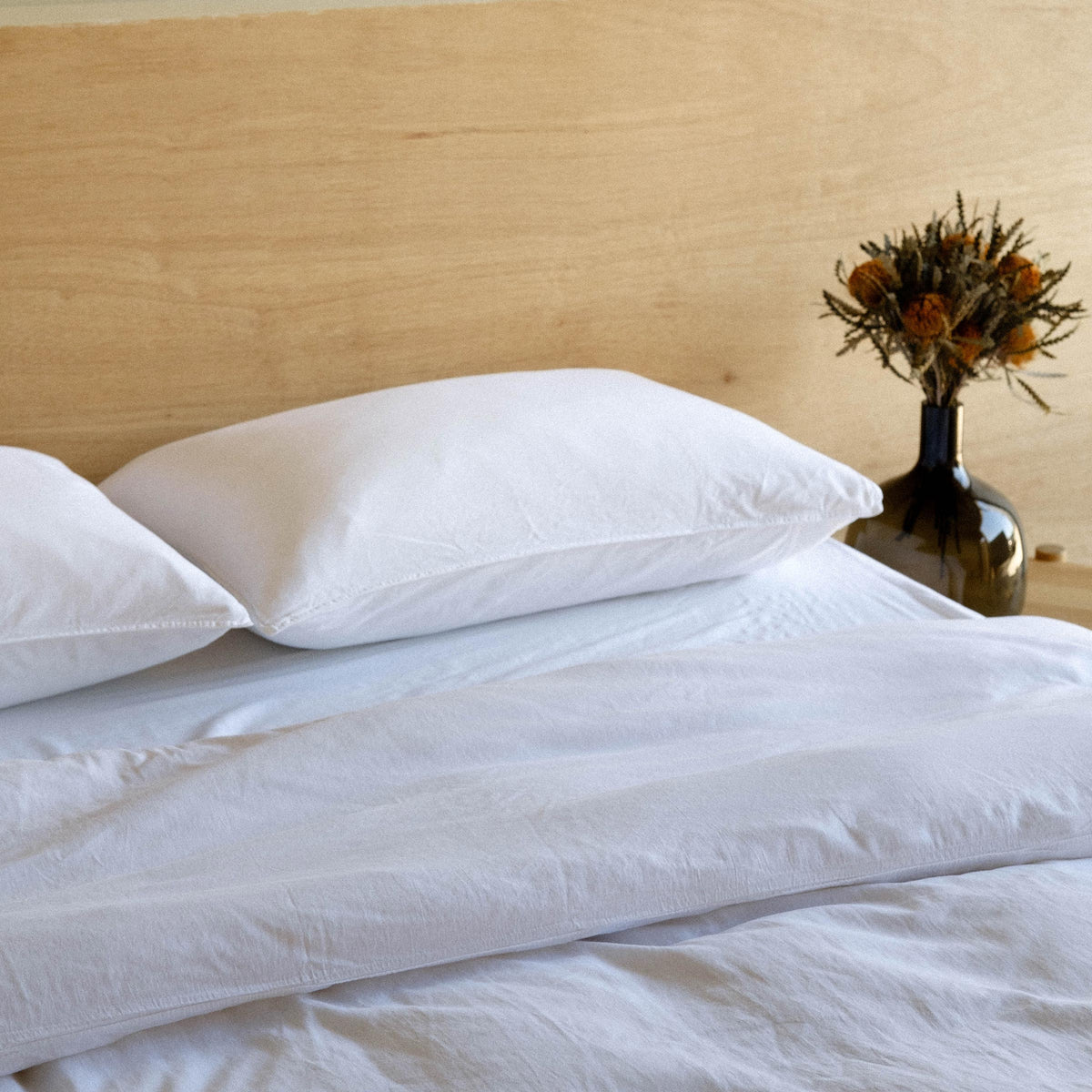 A close up of a bed dressed with white sheets and pillows with a vase of dried flowers 
