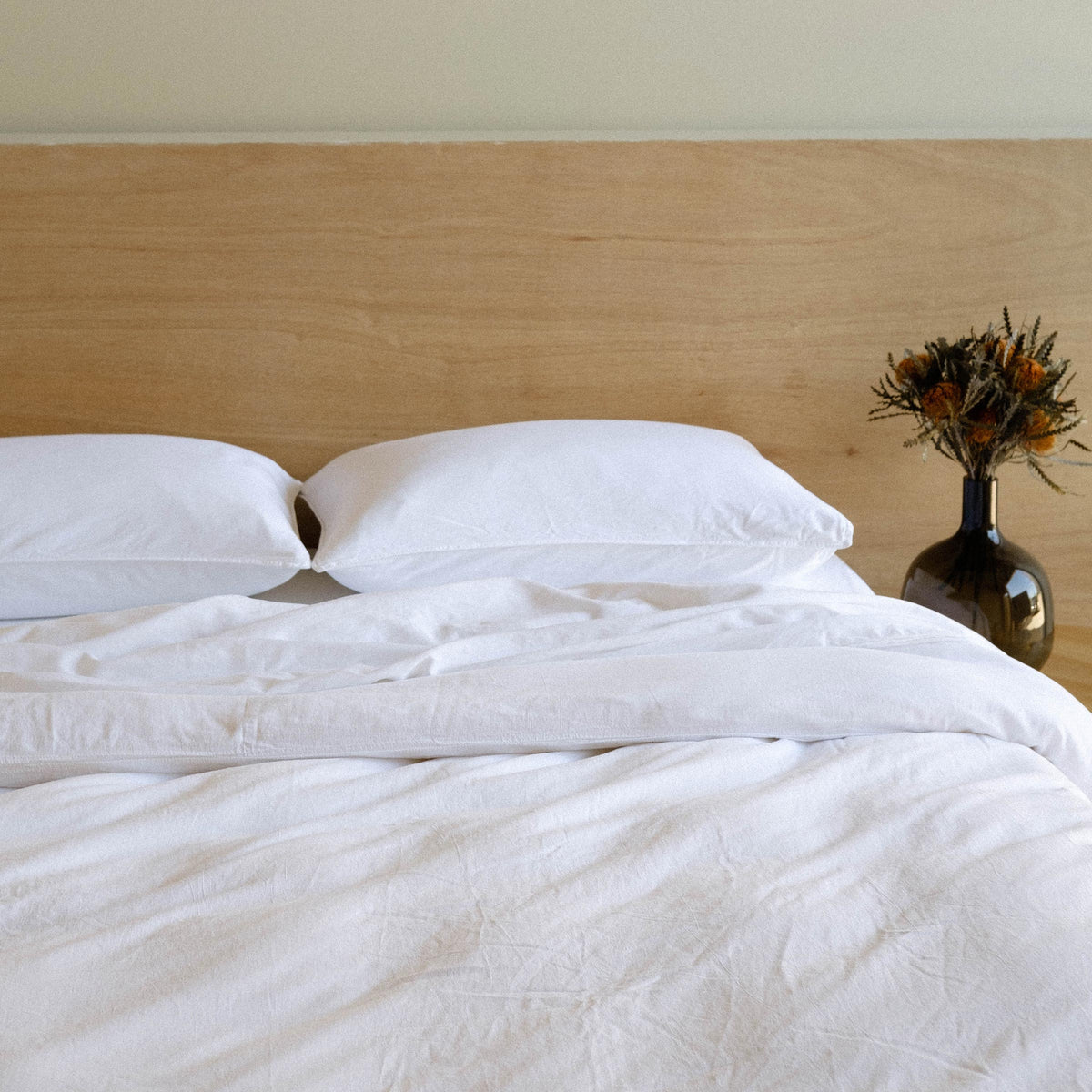 A bed dressed in white matte sateen with two pillows, sheets and a duvet cover in a bedroom setting next to a base of dried flowers